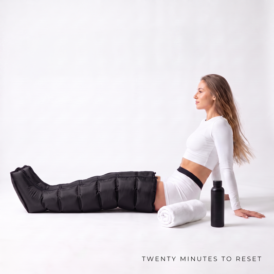 A woman in a white crop top and leggings sits on a white studio floor with a black insulated leg warmer on her extended leg, next to a white towel and a black bottle. The text 'TWENTY MINUTES TO RESET' appears at the bottom.